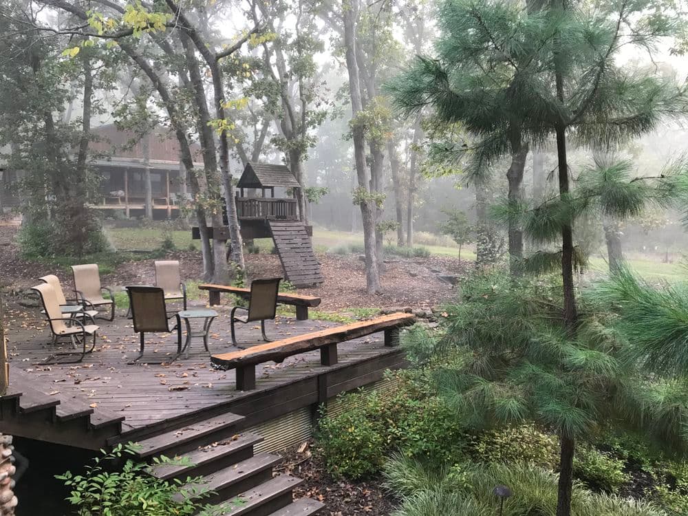 Foggy backyard scene with wooden deck, patio chairs, and playhouse among trees.
