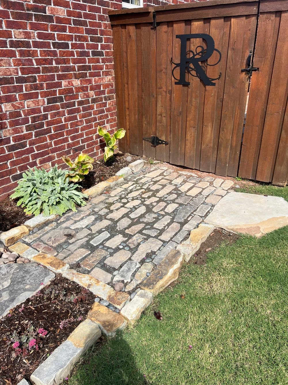 Cobblestone pathway leading to a wooden gate with a decorative letter R, surrounded by plants.