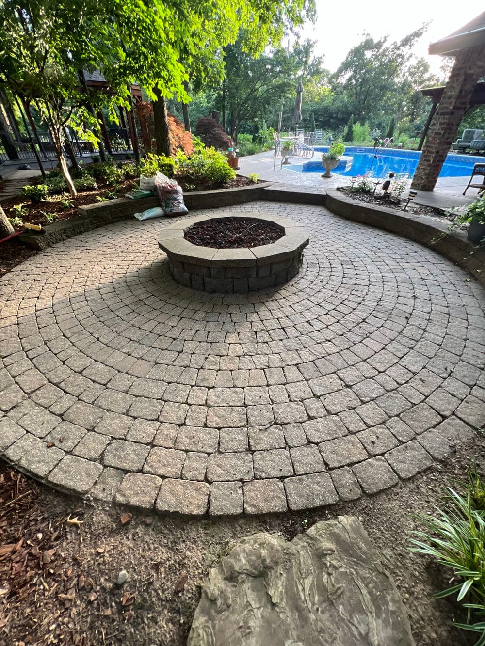 Circular stone fire pit surrounded by landscaped garden and pool area, ideal for outdoor gatherings.